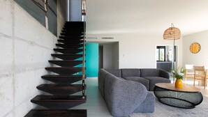 Ein modernes Wohnzimmer mit einer dunklen Holztreppe, einem grauen Sofa, einem runden Couchtisch und einer Korbtischlampe.