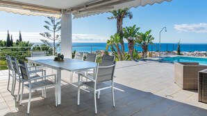 Ein überdachter Patio mit Tisch und Stühlen, mit Blick auf einen Pool und das Meer.