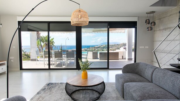 Ein modernes Wohnzimmer mit einem grauen Sofa, einem runden Couchtisch und Meerblick durch große Schiebetüren.