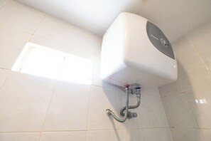 Une salle de bain avec des toilettes blanches, une tuyauterie apparente et une petite fenêtre.