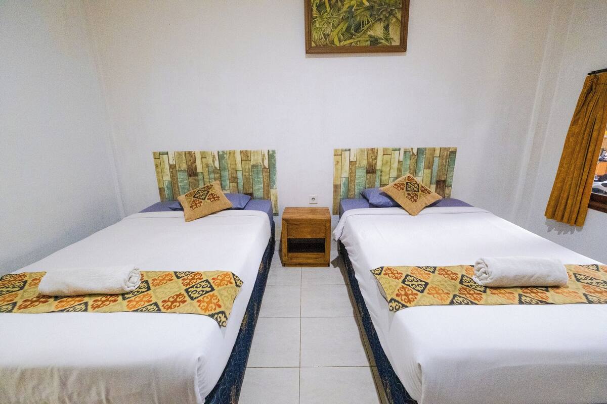 A room with two beds, each with a patterned bedspread and a pillow. There is a small wooden nightstand between the beds.
