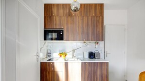 Een kleine keuken met houten kastjes, een Bosch magnetron, een gootsteen, een schaal met fruit, een waterkoker en een broodrooster.