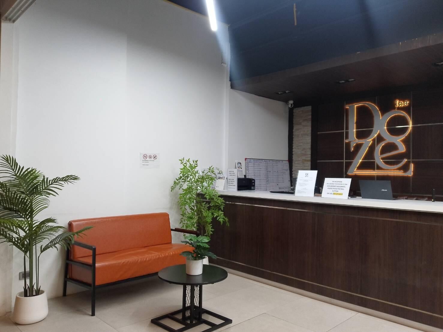A reception area with a dark wooden desk, a neon sign reading "Dez", a sofa, a small round table, and potted plants.