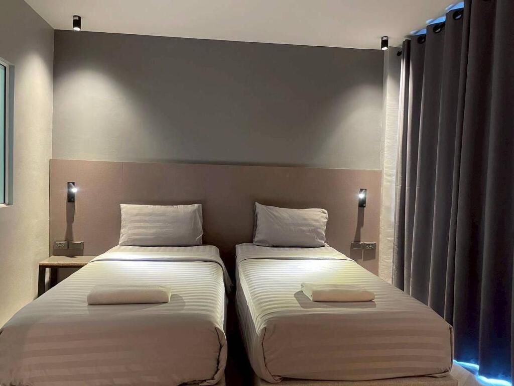 A hotel room with two beds, each with a pillow and a blanket, a nightstand with a lamp, and a window with curtains.