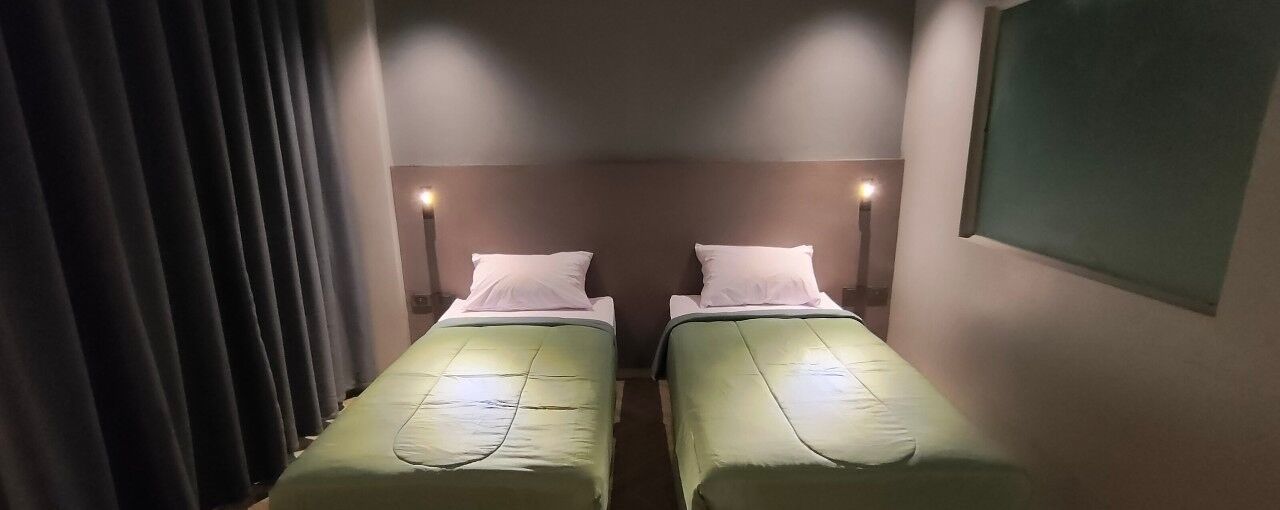 Habitación con dos camas individuales, cada una con una almohada y una colcha verde. Hay luces fijadas en la pared sobre las camas y una ventana al lado derecho.