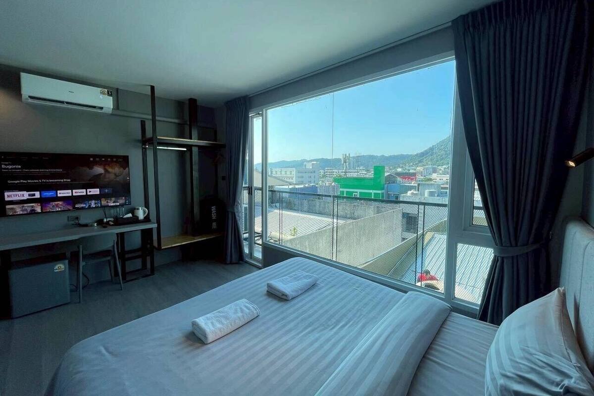 A hotel room with a large bed, a desk with a TV, and a balcony with a city view.
