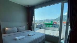 Ein Hotelzimmer mit einem großen Bett, Blick durch eine Schiebetür auf einen Außenbalkon und einer Stadtlandschaft im Hintergrund.