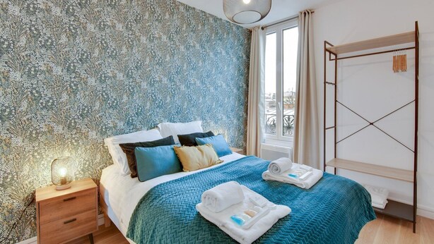 A bedroom with a patterned wallpaper, a bed with multiple pillows, a bedside table with a lamp, a window with curtains, and a wooden shelf unit. - TBS 141 - Bright and elegant apartment (Paris)