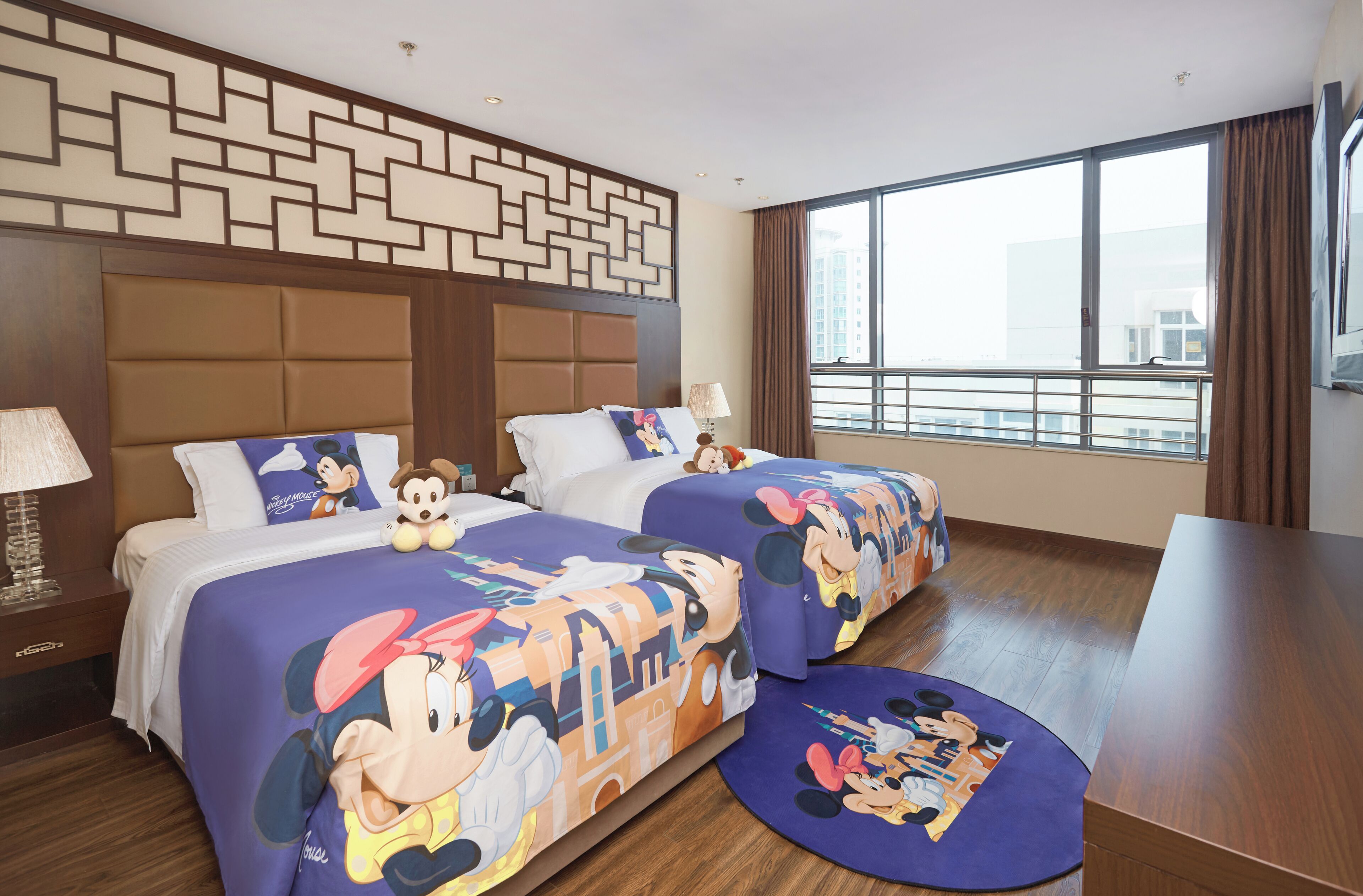 A hotel room with two beds, each covered with Mickey Mouse themed bedding, a large window with a view of buildings, and a wooden floor.