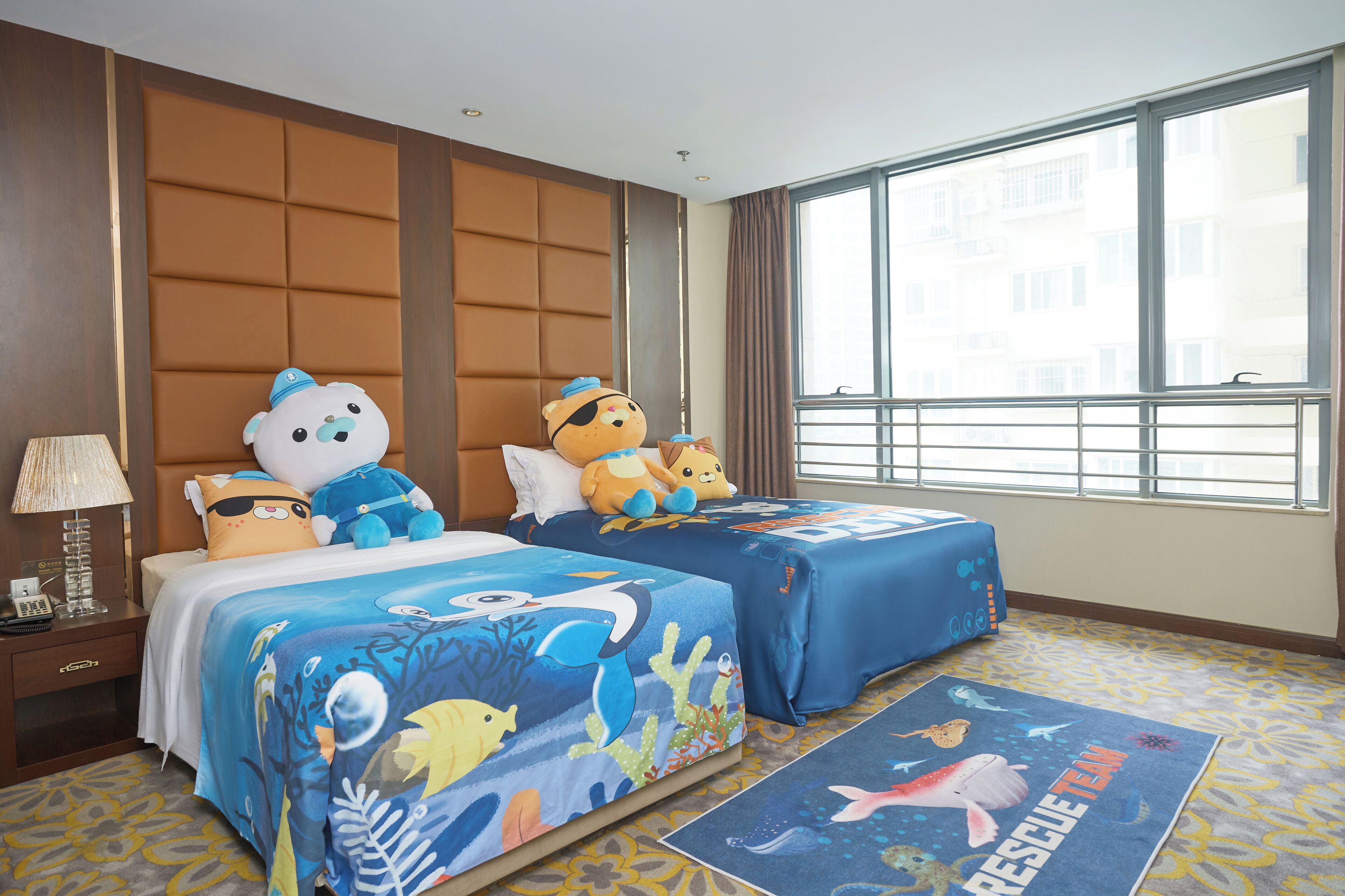 Une chambre d’hôtel avec deux lits, chacun recouvert d’une courtepointe aux motifs marins bleus, une grande fenêtre donnant sur des immeubles et un tapis à motifs sur le sol.