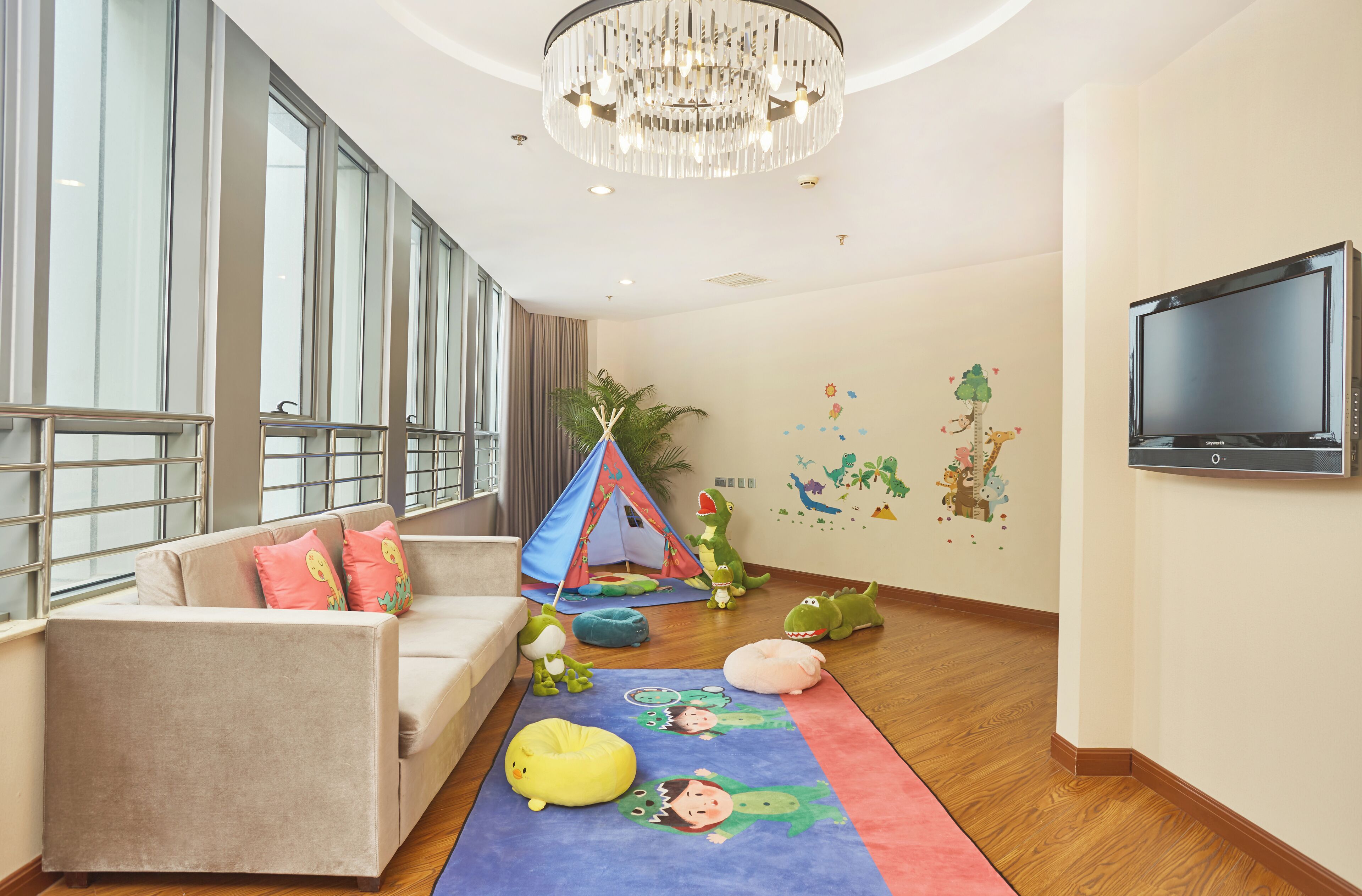 A spacious room with a large window, a beige sofa, a colorful play mat, a tent, and a flat-screen TV mounted on the wall.