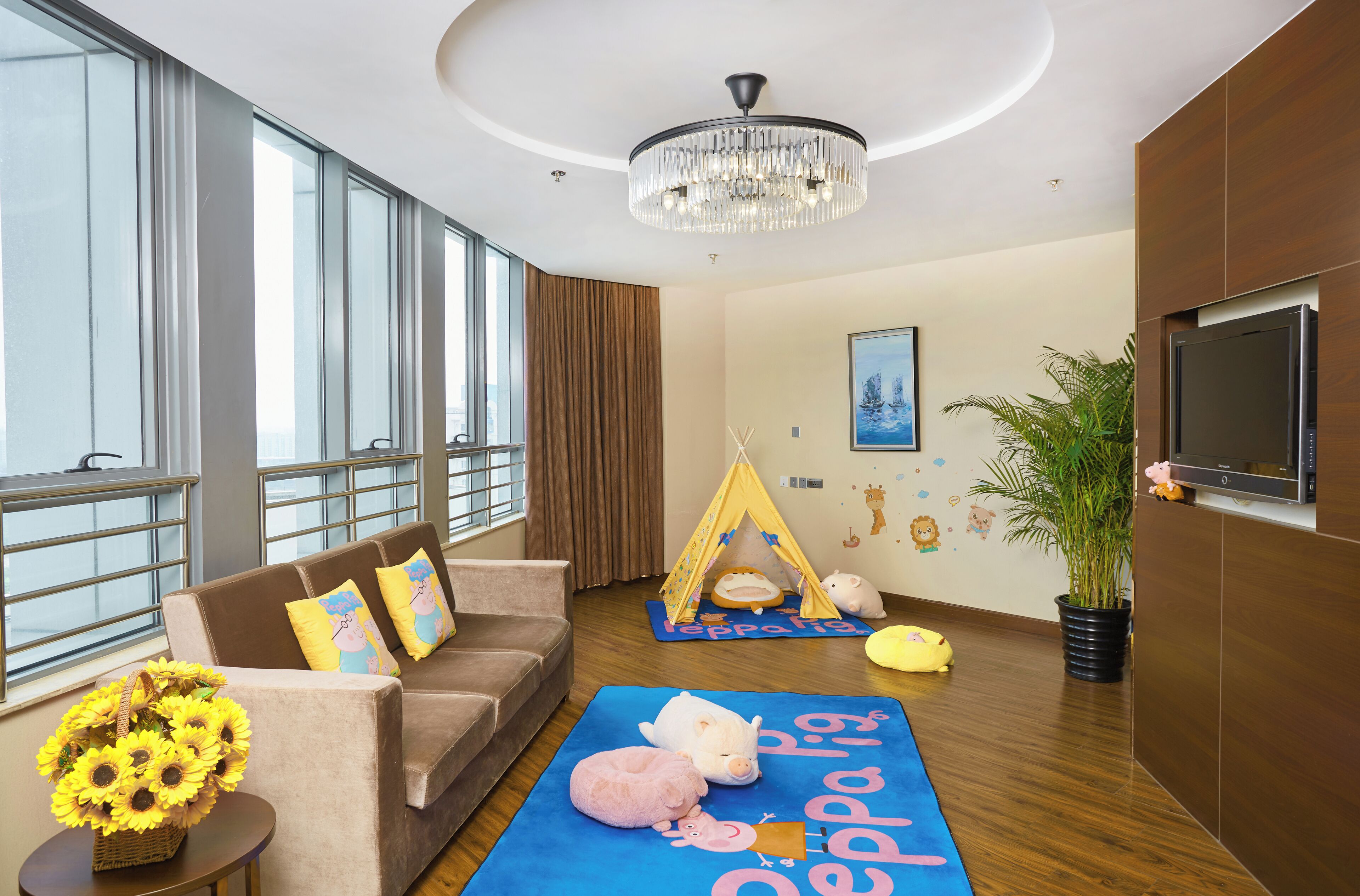 Une chambre d’hôtel moderne dotée d’une grande fenêtre, d’un lustre, d’un canapé avec des coussins, d’un espace de jeux avec une tente, d’une télévision et d’une plante en pot.