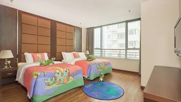 A hotel room with two beds, each with a colorful dinosaur-themed bedspread, a wooden floor, a large window with a view of buildings, and a small rug with a dinosaur design.