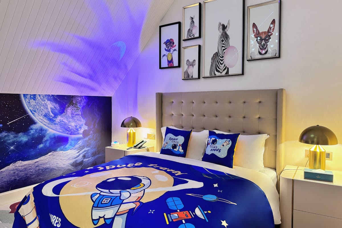 Une chambre à coucher avec une couvre-lit sur le thème de l’espace, une tête de lit capitonnée et des œuvres d’art encadrées accrochées au mur.