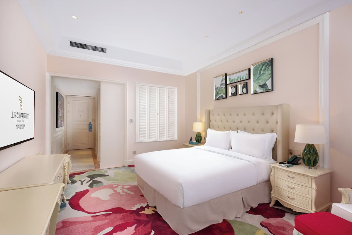 Une chambre d’hôtel avec un grand lit, une table de chevet avec une lampe, une petite table de chevet avec un téléphone, un banc rouge et une moquette à motifs floraux.