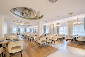 Een ruime eetruimte met meerdere tafels en stoelen, grote ramen en stijlvolle kroonluchters.