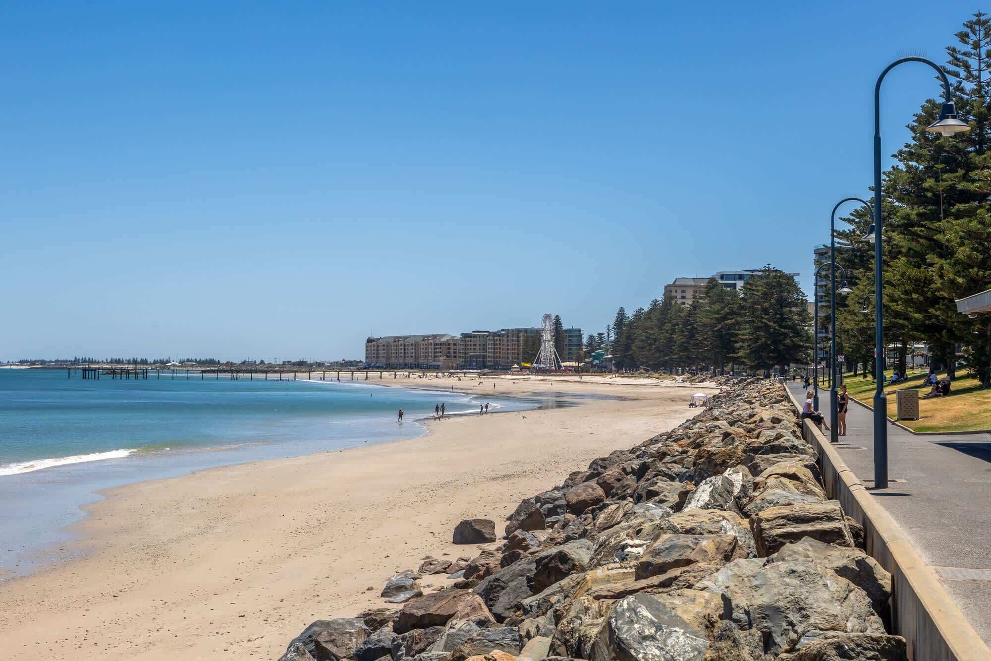 A beach with a rocky breakwater, clear blue water, and buildings in the background.
