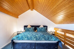Ein Schlafzimmer mit schrägen Wänden, einem Bett mit gemustertem Überwurf, zwei Nachttischen mit Lampen und einem Holz-Kopfteil.