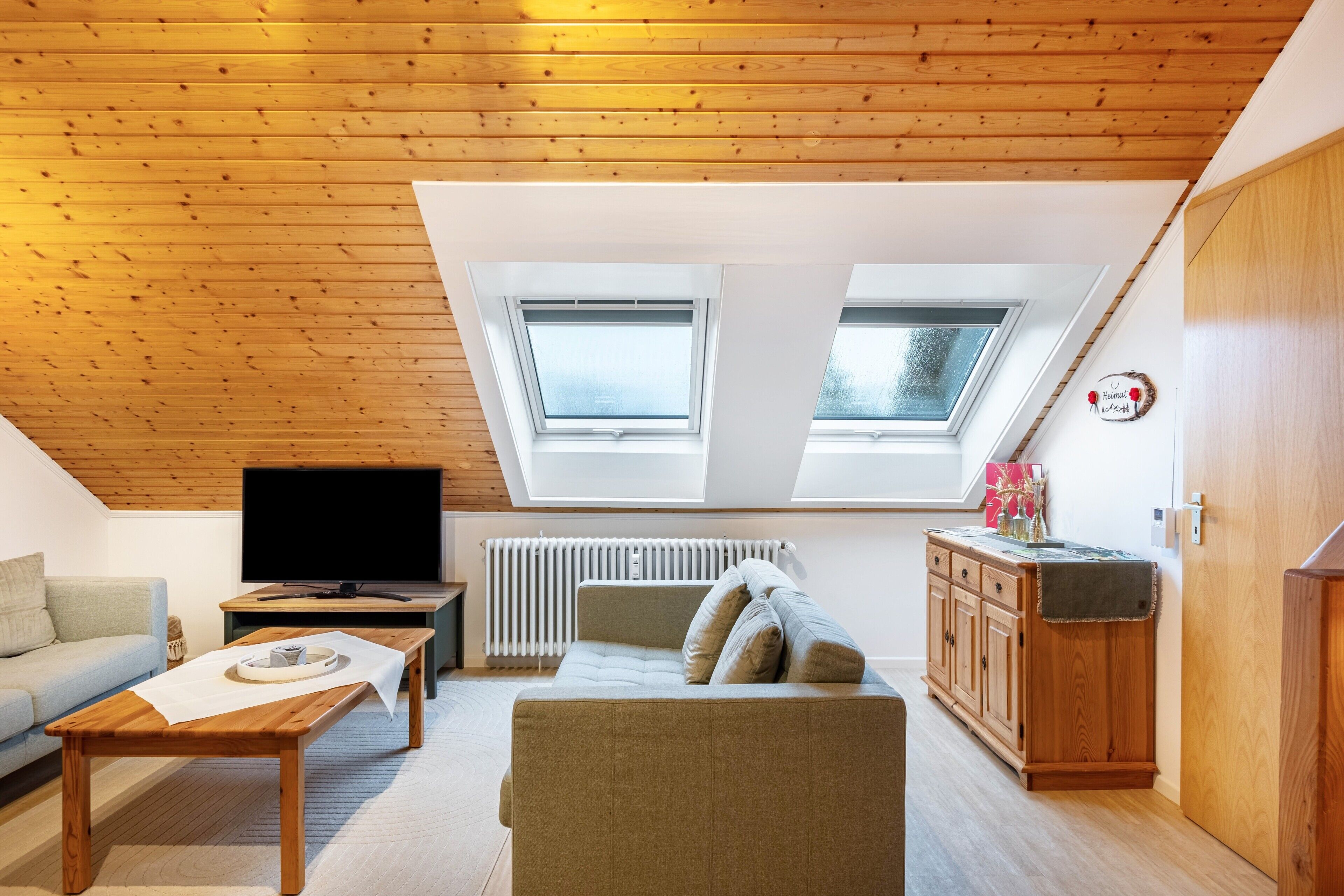 Ein Loft-Zimmer mit Holzbalkendecke, zwei Dachfenstern, einem Fernseher, einem Sofa, einem Couchtisch und einem Schrank.
