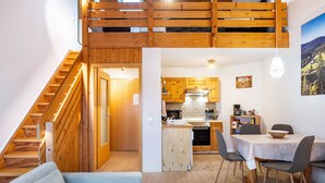 A loft-style room with a wooden staircase, a dining area with a table and chairs, and a kitchen with wooden cabinets and a hanging light fixture.