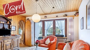 Ein gemütliches Zimmer mit Holzbalkendecke, einem Fernseher, einer Couch und einem Tisch mit Blumen.