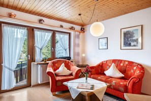 A living room with a wooden ceiling, a red patterned sofa, a red armchair, a table with a vase, and a window with curtains.