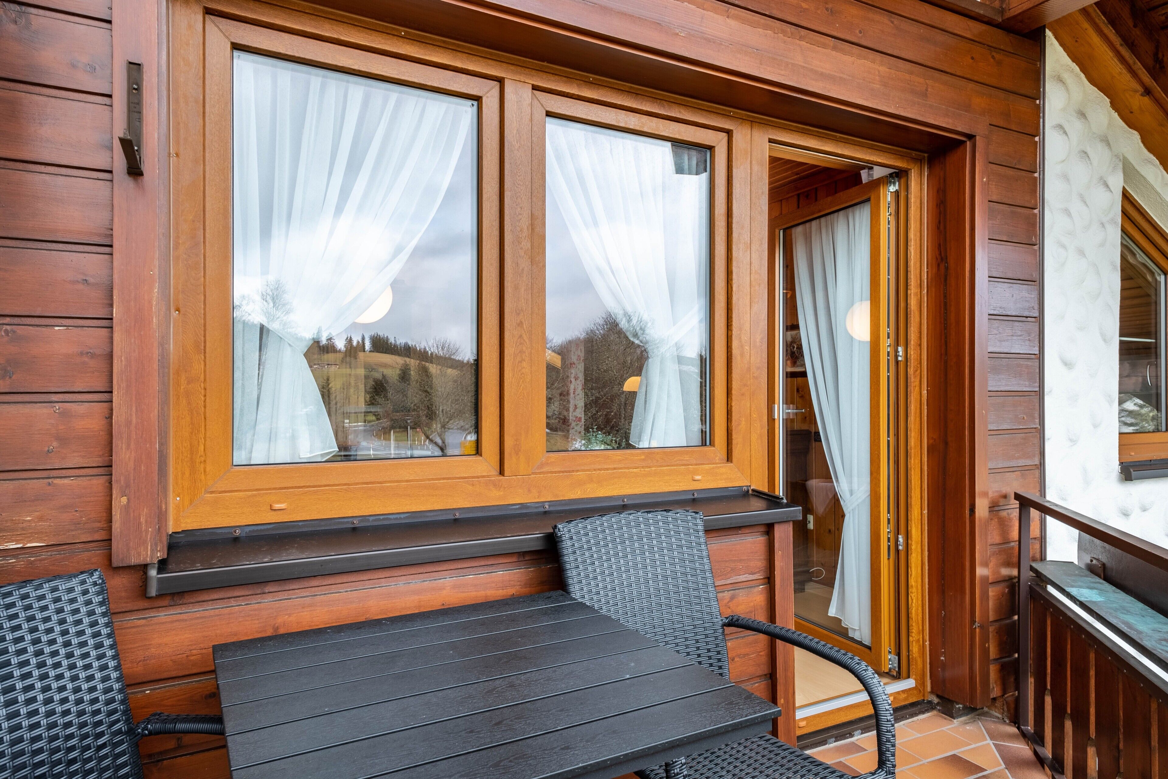 Eine Holzhütte mit Tisch und Stühlen davor, großen Fenstern mit Vorhängen und einer Holztür.