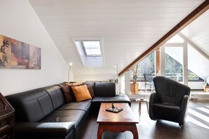 A modern living room with a black leather sofa, a wooden coffee table, and a black armchair. There is a skylight above the sofa and large windows providing natural light.