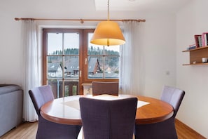 Un coin repas comprenant une table ronde, quatre chaises, une lampe suspendue, de grandes fenêtres avec des rideaux et une bibliothèque.