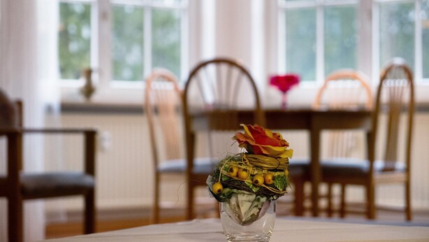 Une table à manger avec des chaises, un vase orné d’un arrangement floral et une fenêtre donnant sur des arbres.