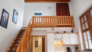 A staircase leading to a loft bedroom with a wooden railing, a kitchen area with white cabinets, and a window with blinds.