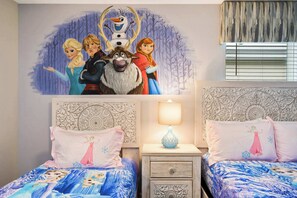 Une chambre avec deux lits, chacun agrémenté d’une couvre-lit à motifs, une table de chevet avec une lampe et une fresque murale représentant des personnages de dessins animés.