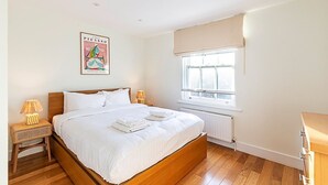 A bedroom with a wooden bed, white bedding, a window with beige curtains, a radiator, and a poster on the wall. - Stylish Chelsea Flat with Luxe Comfort & Charm (London)
