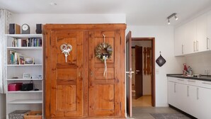 Une armoire en bois à deux portes, une bibliothèque remplie de livres et d’objets décoratifs, et un coin cuisine avec des placards et un plan de travail.
