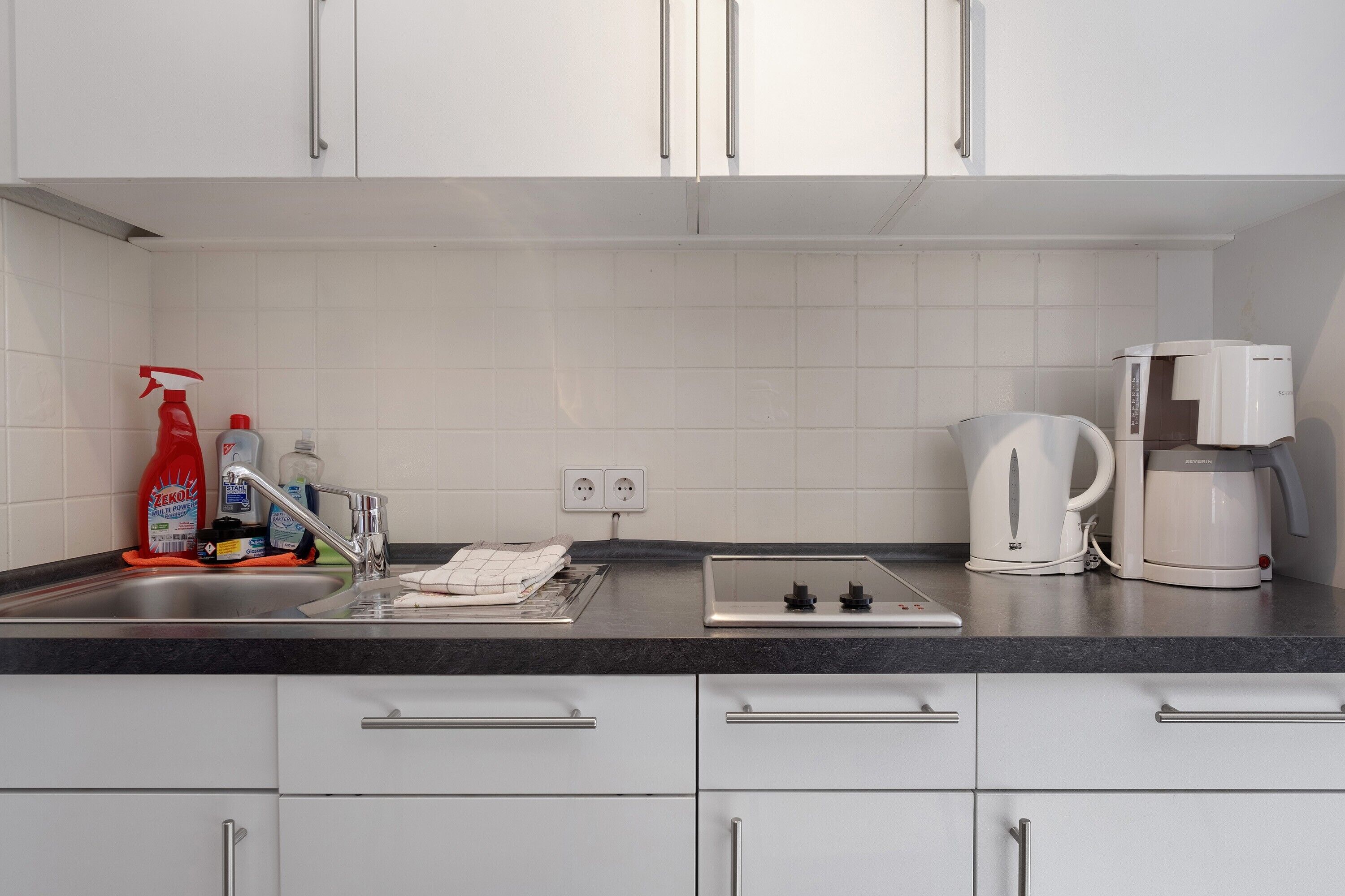 A modern kitchen with white cabinets, a stainless steel sink, a kettle, and a coffee maker.
