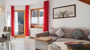 A living room with a sofa, a coffee table, a dining table with chairs, a radiator, and a window with red curtains.