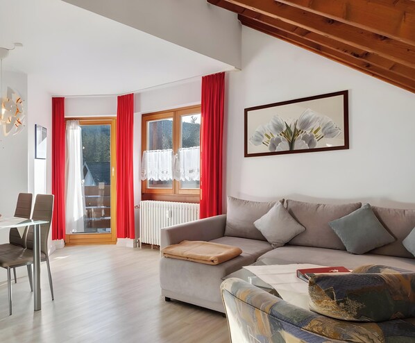 Una sala de estar con un sofá, una mesa de centro, una mesa de comedor con sillas, un radiador y una ventana con cortinas rojas.
