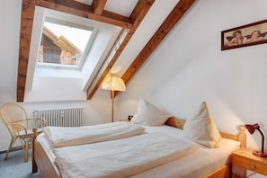 Une chambre avec un plafond en pente, une fenêtre, un radiateur, un lit avec des oreillers, une chaise et une table de chevet avec une lampe.