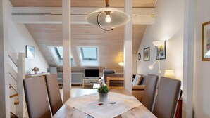 Un coin repas comprenant une table, des chaises, un luminaire suspendu et donnant sur un salon avec un canapé et un téléviseur.