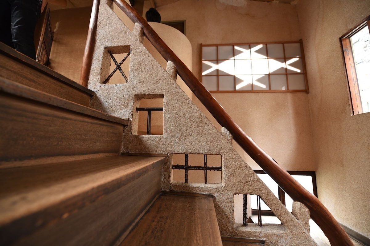 A wooden staircase with a handrail, leading to a room with a window featuring a geometric pattern.