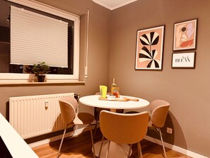 A dining area with a round table, four chairs, a potted plant, a radiator, and two framed artworks on the wall.