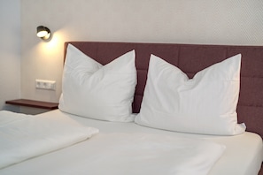 A neatly made bed with white bedding and two white pillows, a brown upholstered headboard, and a wall-mounted light fixture.