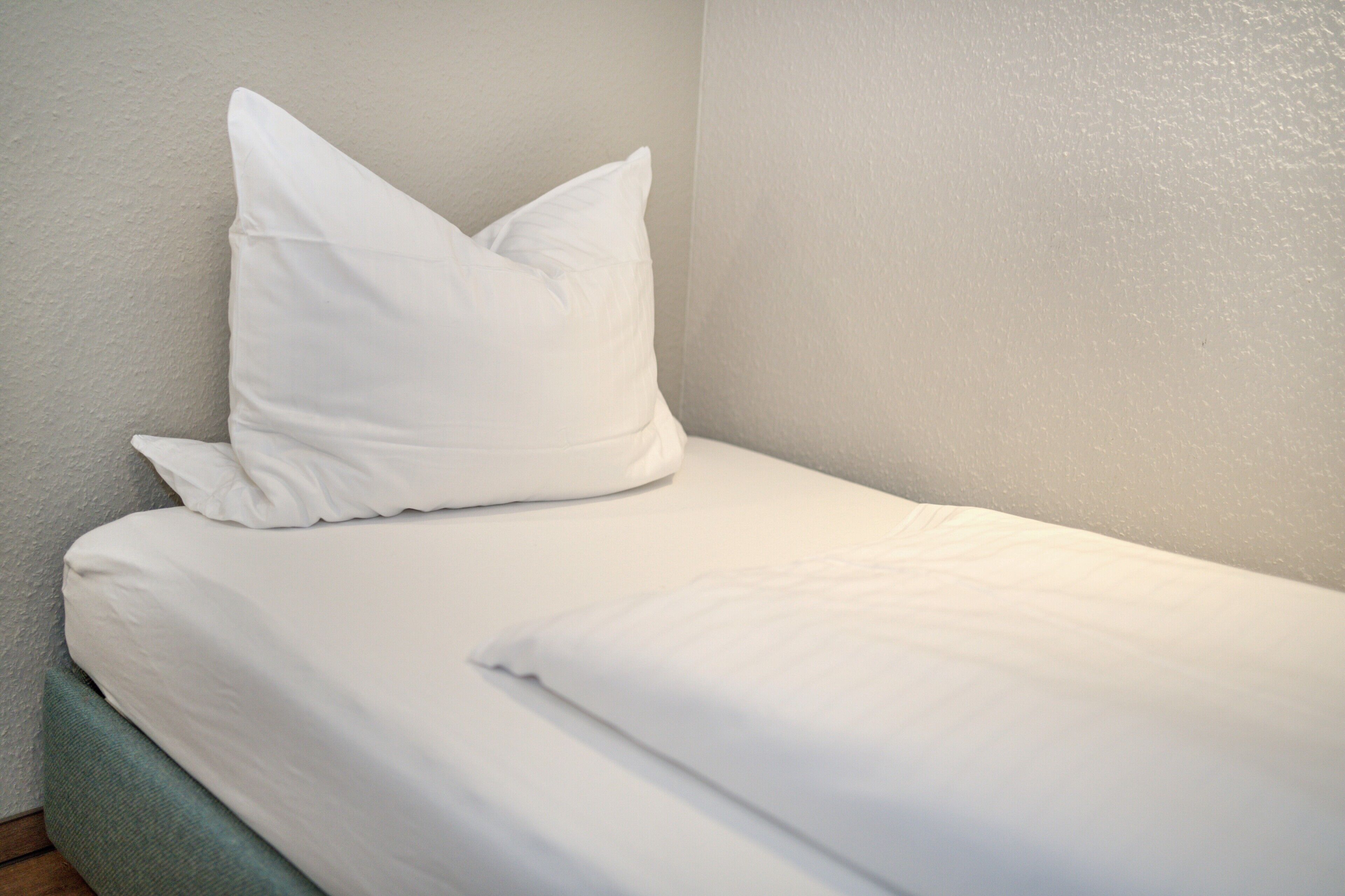 Un lit simple avec une literie et des oreillers blancs, un mur uni et un plancher en bois.