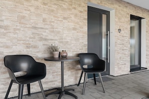 Une façade moderne avec un mur en pierre, deux chaises noires, une petite table ronde et une porte.