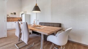 A modern dining area with a wooden table, white and grey chairs, a black pendant light, and a kitchen in the background.