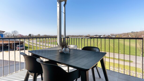 Uma varanda com mesa e cadeiras, vista para uma estrada e um edifício ao fundo.