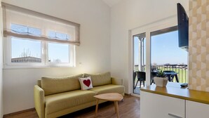 Quarto com sofá, uma pequena mesa de madeira, televisão e vista através de uma porta de vidro.