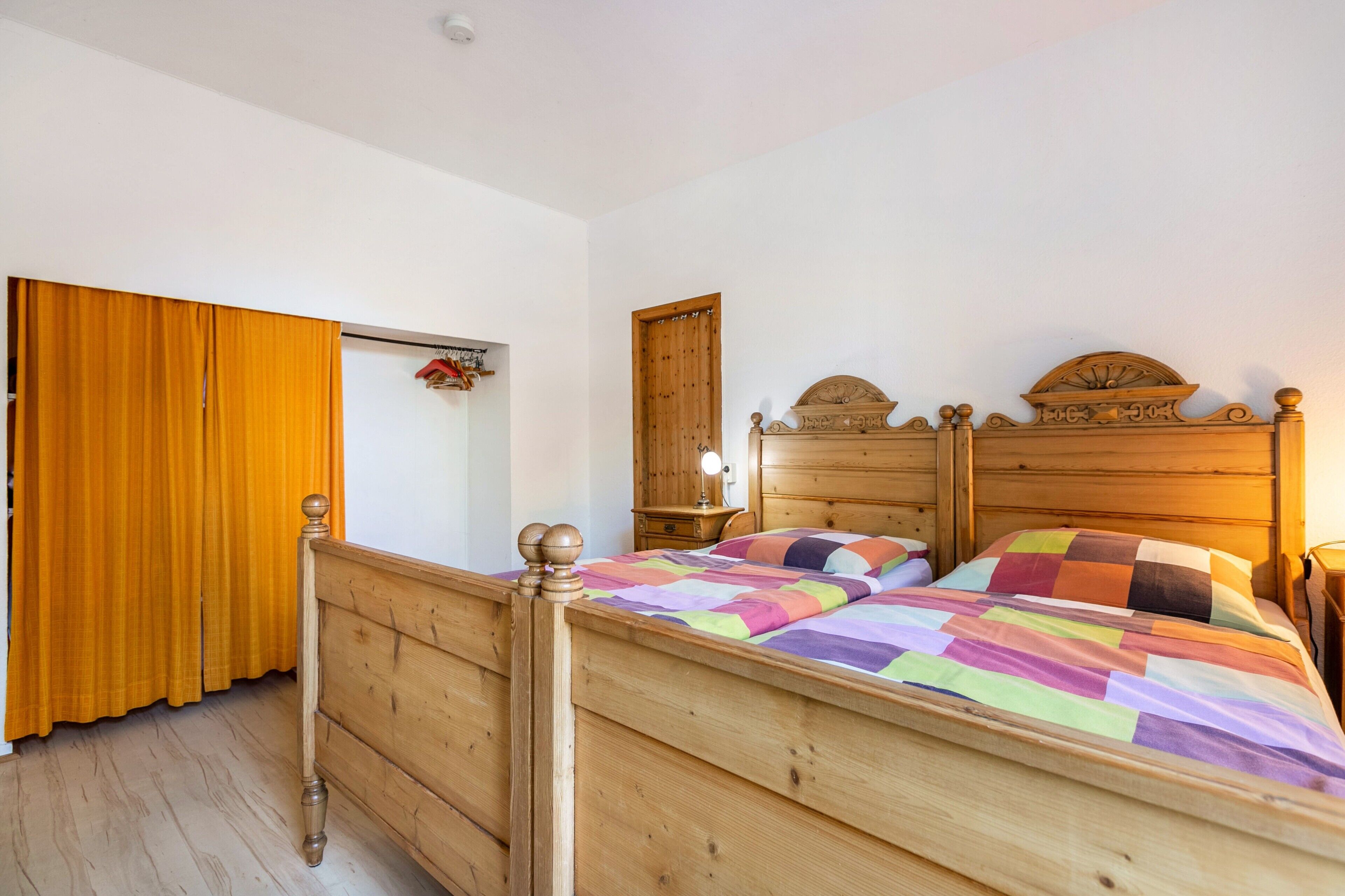 Un lit en bois avec une couverture colorée, une tête de lit en bois, une table de chevet en bois et une armoire en bois avec un rideau.