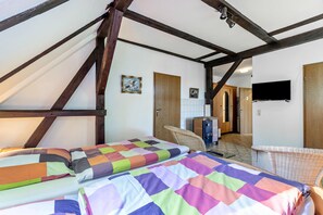 Ein Zimmer mit schrägem Dach, Holzbalken, einem Bett mit farbenfroher Steudecke, einem an der Wand montierten Fernseher und einem kleinen Schreibtisch mit Stuhl.