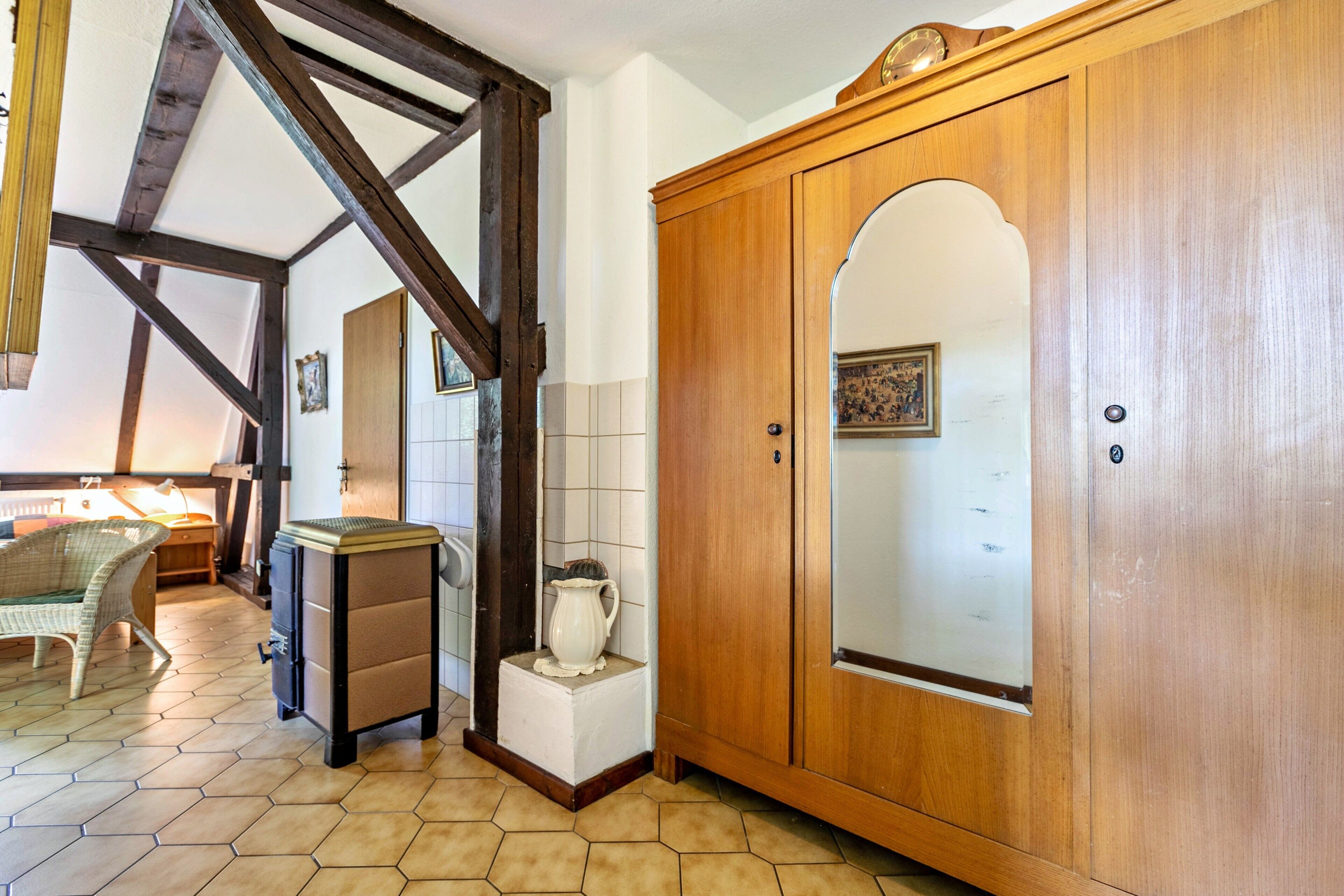 A room with a wooden wardrobe, a wicker chair, a small cabinet, and a tiled floor.